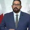Man in a blue suit giving a speech at a press conference with the Mexican flag and FGGR banner in the background.