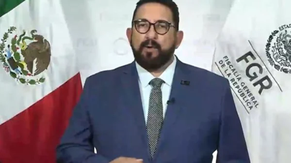 Man in a blue suit giving a speech at a press conference with the Mexican flag and FGGR banner in the background.