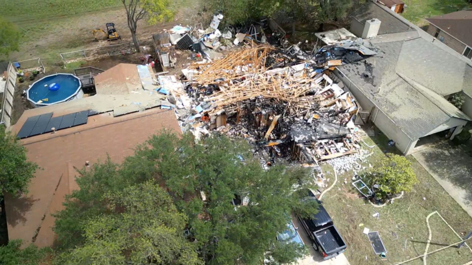Aerial view of a collapsed house with tangled beams, rubble and a damaged roof; a round blue pool and yard are visible to the left.