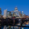 City skyline at dusk with tall glass buildings and a graffiti-covered bridge over a calm river, reflecting city lights.