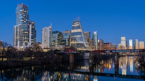 City skyline at dusk with tall glass buildings and a graffiti-covered bridge over a calm river, reflecting city lights.