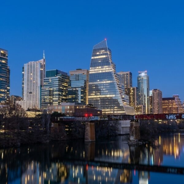 City skyline at dusk with tall glass buildings and a graffiti-covered bridge over a calm river, reflecting city lights.