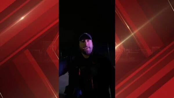 Video still of a man in a cap speaking to the camera at night, dark background.