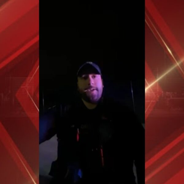 Video still of a man in a cap speaking to the camera at night, dark background.