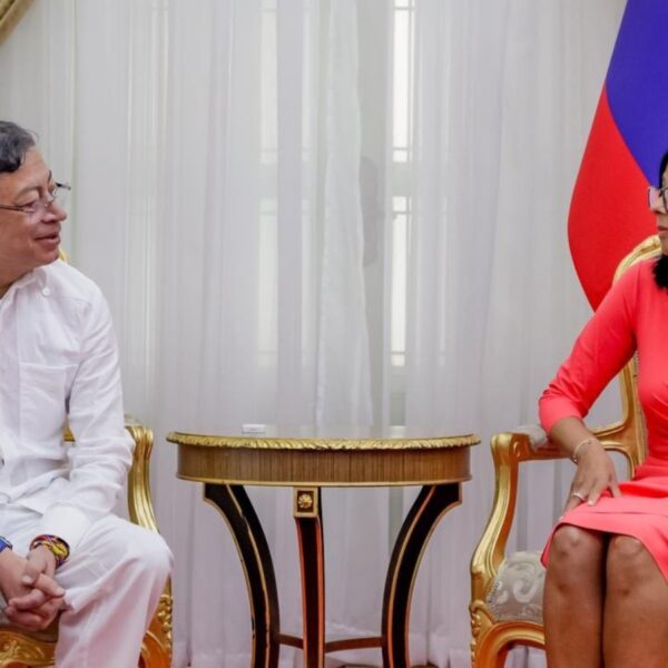 Two officials sit in ornate gold chairs facing each other in a formal room, with flags in the background and a small round table between them.