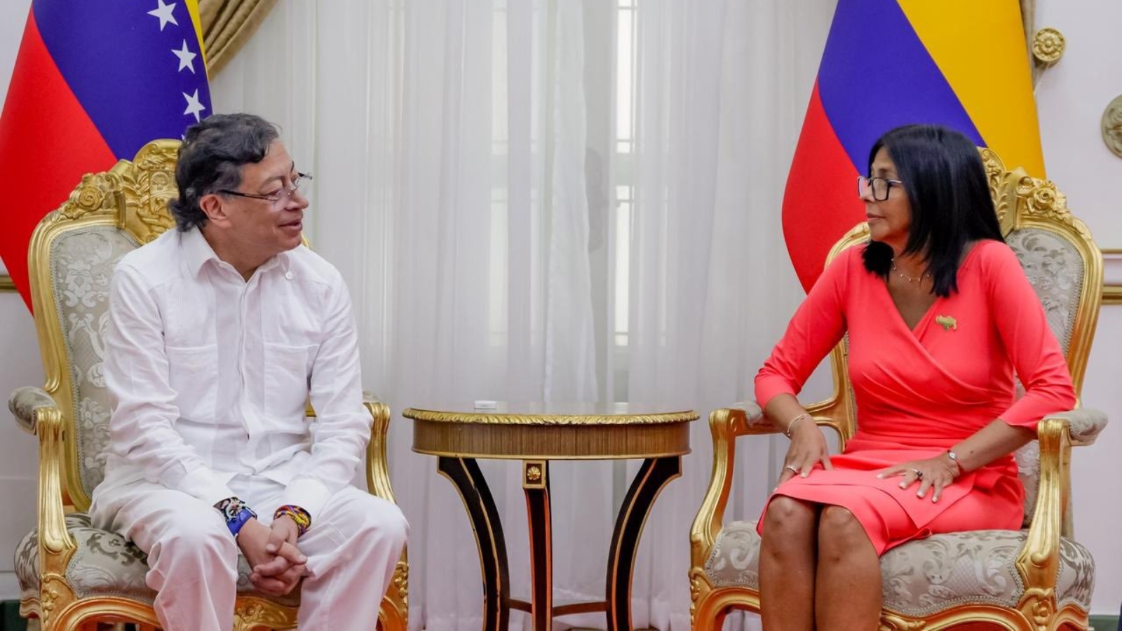 Two officials sit in ornate gold chairs facing each other in a formal room, with flags in the background and a small round table between them.