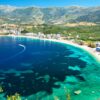 Aerial view of a turquoise bay with a curved sandy beach and a town along the shoreline, backed by green mountains.