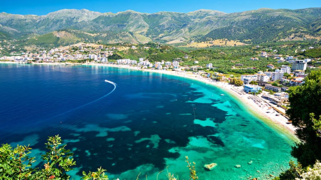 Aerial view of a turquoise bay with a curved sandy beach and a town along the shoreline, backed by green mountains.