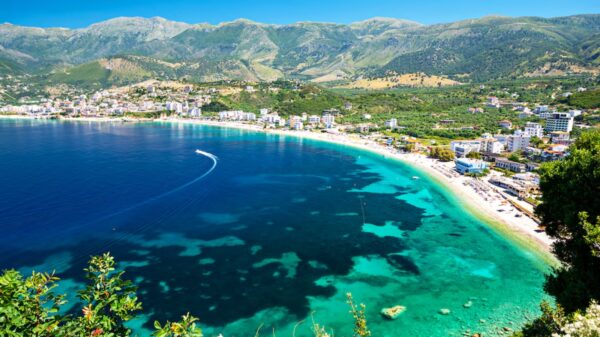Aerial view of a turquoise bay with a curved sandy beach and a town along the shoreline, backed by green mountains.