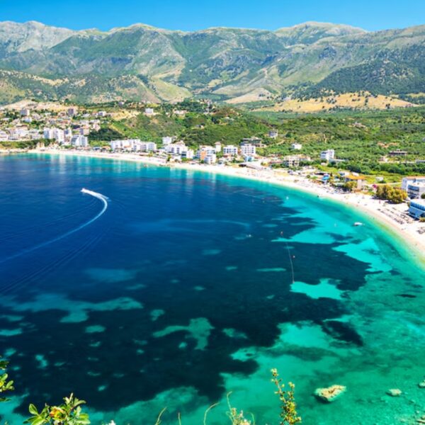 Aerial view of a turquoise bay with a curved sandy beach and a town along the shoreline, backed by green mountains.