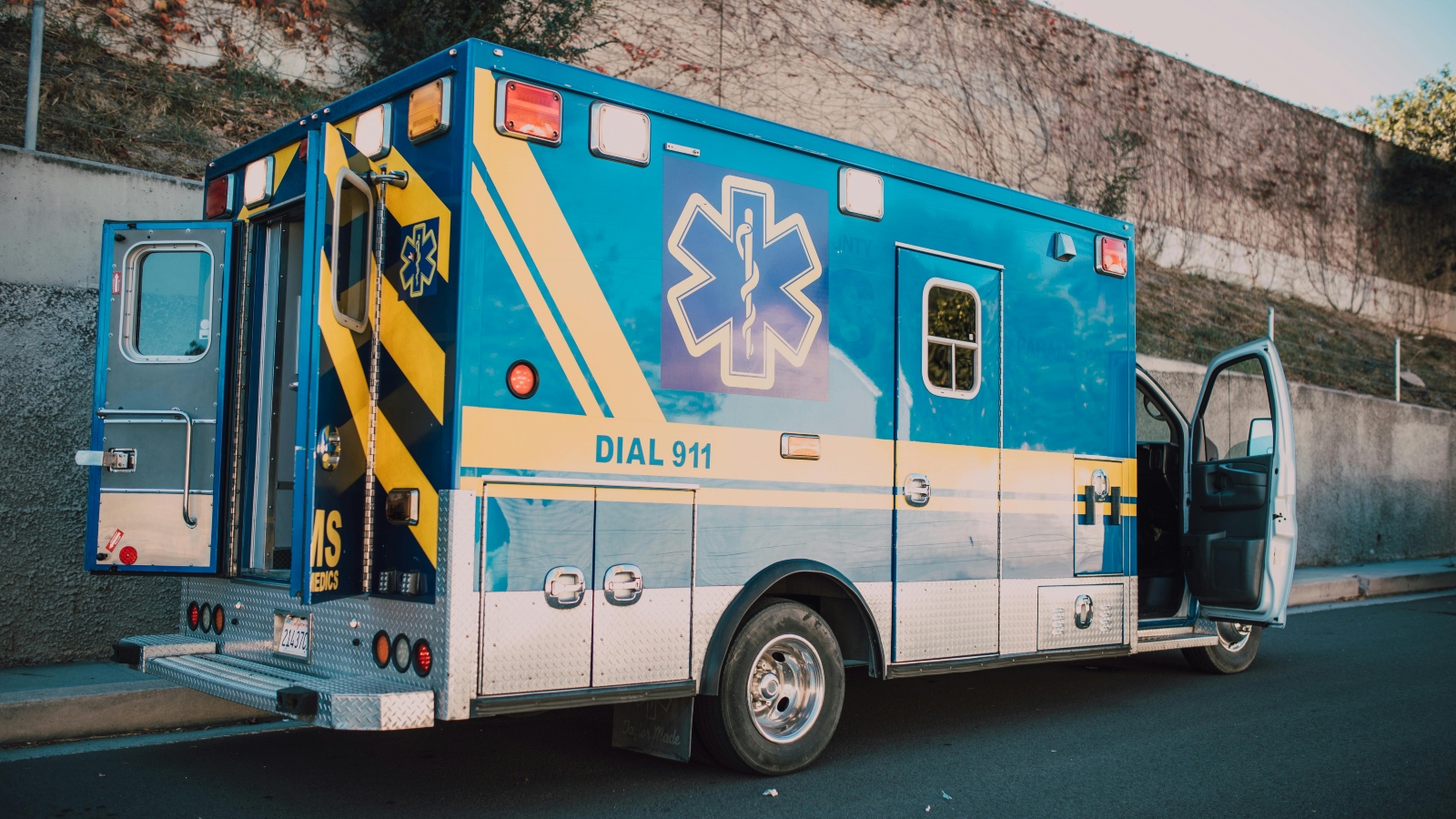 Blue emergency ambulance parked on a street with the side door open, showing 'DIAL 911' on the side panel and medical insignia visible.