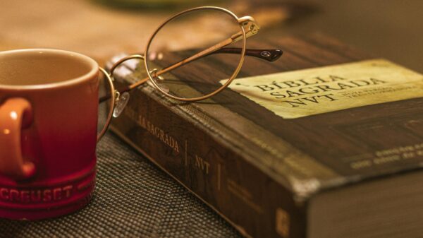 Round gold-rimmed glasses rest on an old Bible beside a red mug, creating a warm, cozy reading scene. On the Bible’s cover and page edges you can see weathered typography and a Spanish Bible title, La Biblia Sagrada NT.
