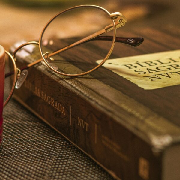 Round gold-rimmed glasses rest on an old Bible beside a red mug, creating a warm, cozy reading scene. On the Bible’s cover and page edges you can see weathered typography and a Spanish Bible title, La Biblia Sagrada NT.