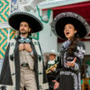 Two mariachi performers in traditional outfits, singing and posing on a colorful festival street.