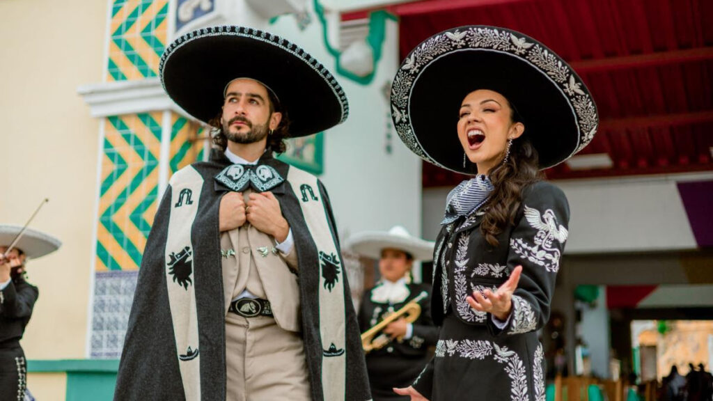 Two mariachi performers in traditional outfits, singing and posing on a colorful festival street.
