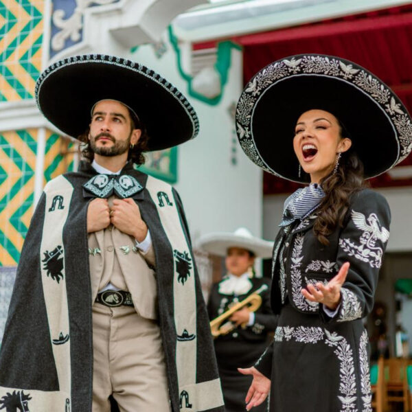 Two mariachi performers in traditional outfits, singing and posing on a colorful festival street.