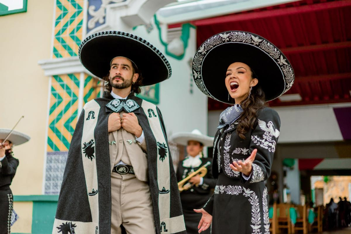 Two mariachi performers in traditional outfits, singing and posing on a colorful festival street.