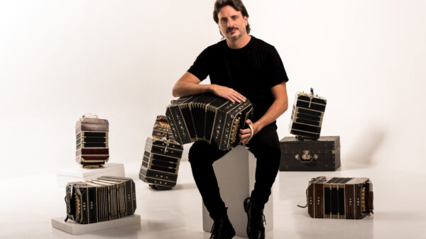 Man in a black shirt seated on a white block, playing a large black accordion in a studio surrounded by several vintage accordions and cases.