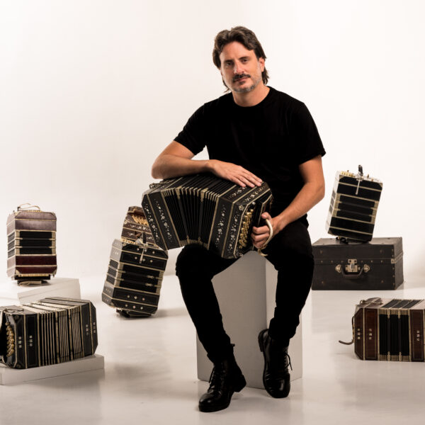 Man in a black shirt seated on a white block, playing a large black accordion in a studio surrounded by several vintage accordions and cases.