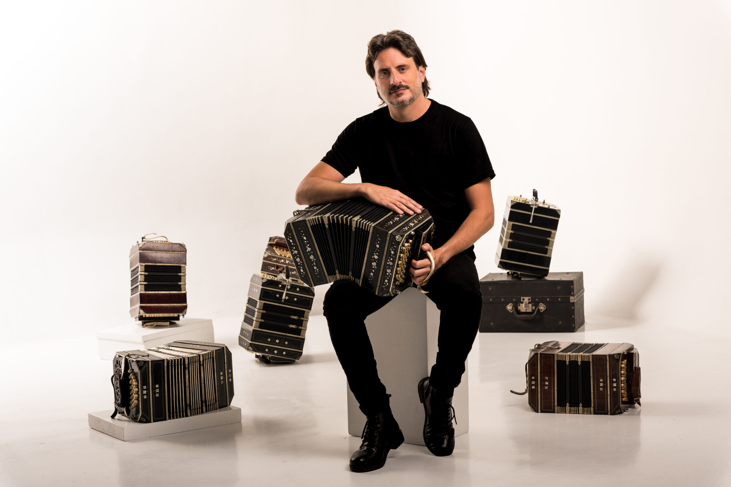 Man in a black shirt seated on a white block, playing a large black accordion in a studio surrounded by several vintage accordions and cases.