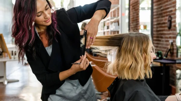 Hairdresser trims a client's blonde shoulder-length hair with a comb and scissors in a bright salon.