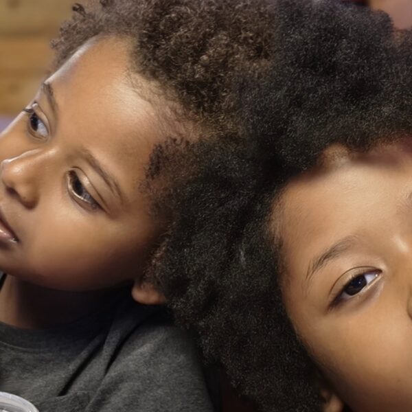 Two children with textured hair leaning their heads together, one holding a drink with a straw visible in the foreground.