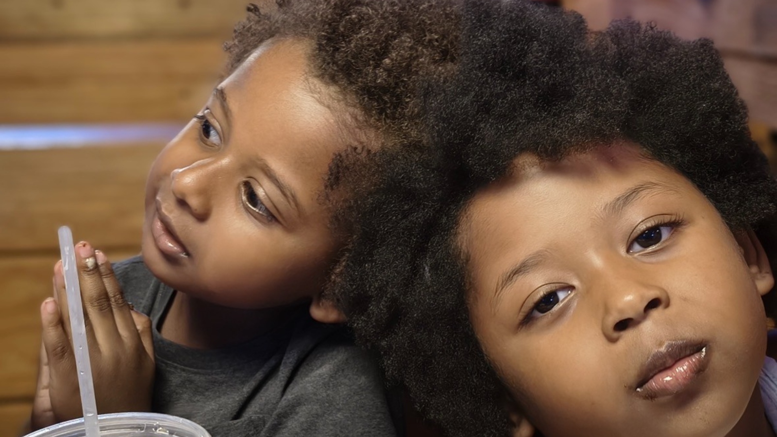Two children with textured hair leaning their heads together, one holding a drink with a straw visible in the foreground.