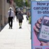 Outdoor ad promoting in-store payment in 4 installments, showing a hand holding a smartphone with a card app on the right; blurred shoppers in the background.