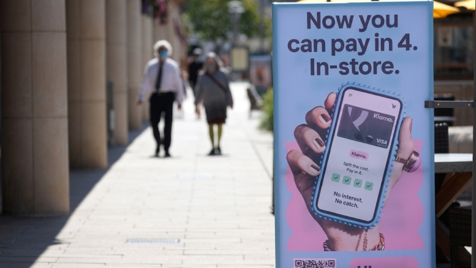 Outdoor ad promoting in-store payment in 4 installments, showing a hand holding a smartphone with a card app on the right; blurred shoppers in the background.