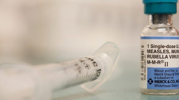 Close-up of a vaccine vial labeled MMR II and a syringe on a clinical surface, blue cap visible in the background.
