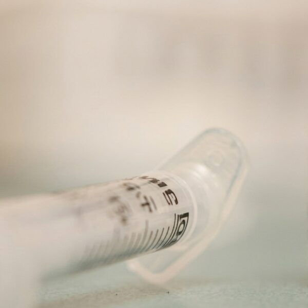Close-up of a vaccine vial labeled MMR II and a syringe on a clinical surface, blue cap visible in the background.