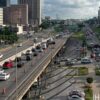 Busy urban highway with multiple lanes and orange construction cones; surrounding office buildings and greenery nearby.