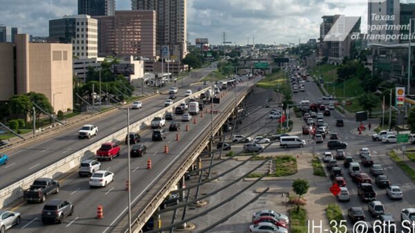 Busy urban highway with multiple lanes and orange construction cones; surrounding office buildings and greenery nearby.