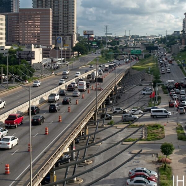 Busy urban highway with multiple lanes and orange construction cones; surrounding office buildings and greenery nearby.