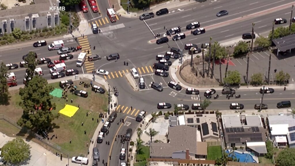 Aerial view of a busy intersection with many police cars and a police presence, plus people gathered in a park with yellow and green tents nearby.