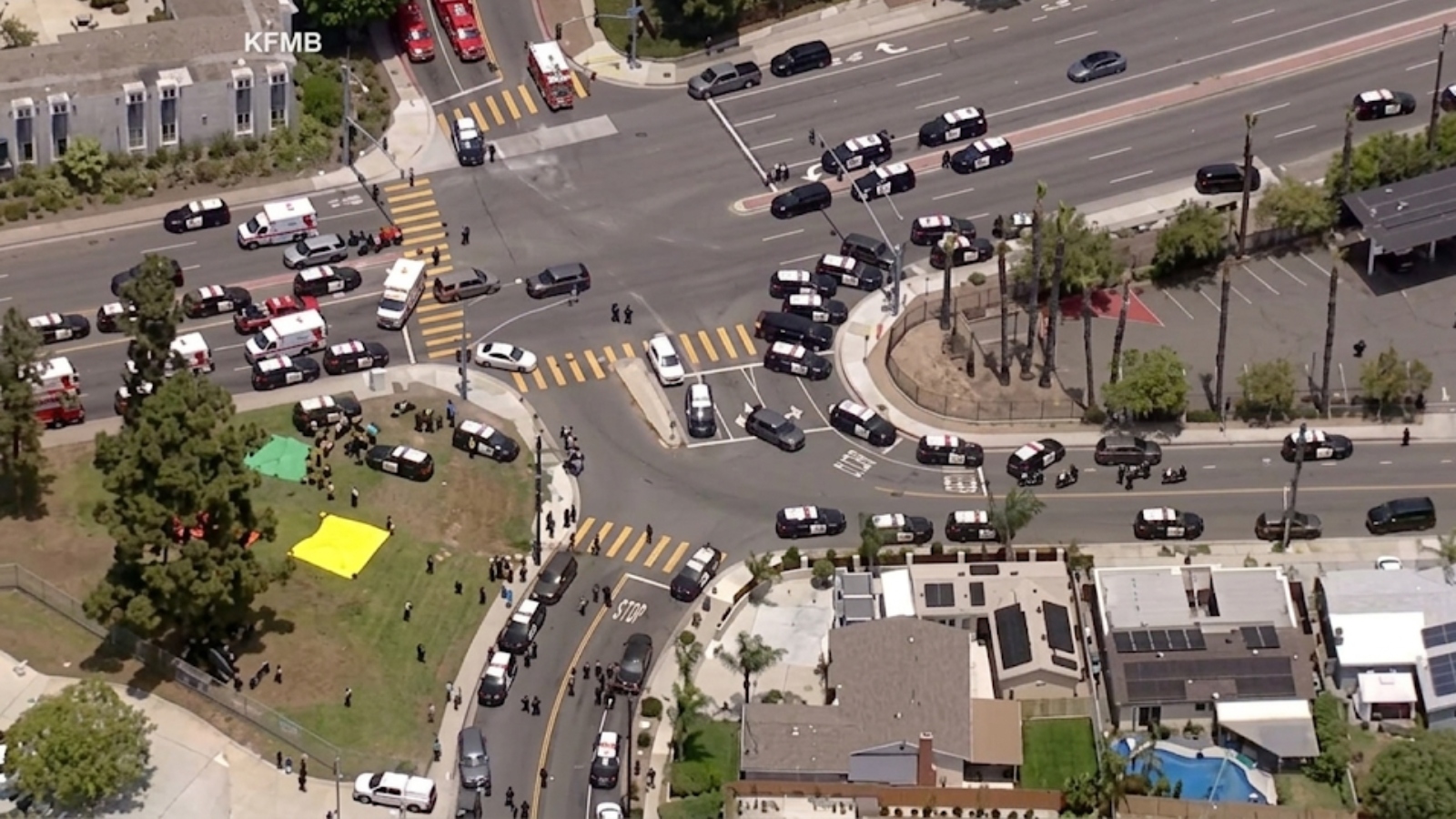 Aerial view of a busy intersection with many police cars and a police presence, plus people gathered in a park with yellow and green tents nearby.