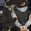 Police officers in tactical vests escort a handcuffed man; patches read 'POLICE ICE' on their backs.
