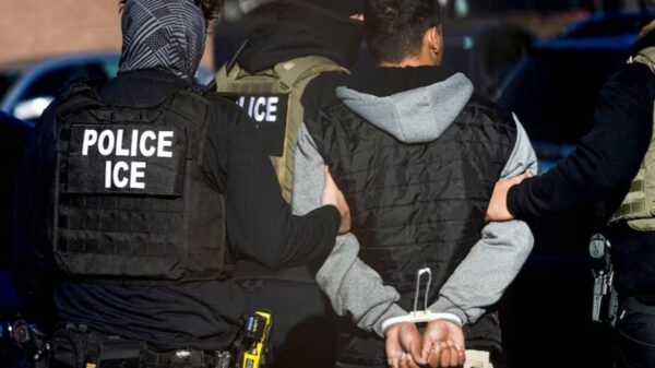Police officers in tactical vests escort a handcuffed man; patches read 'POLICE ICE' on their backs.