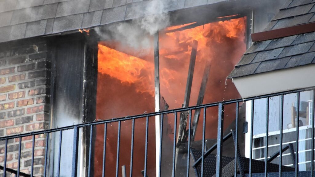 Balcony doorway on fire with bright flames and rising smoke, metal railing in front and brick wall to the left.