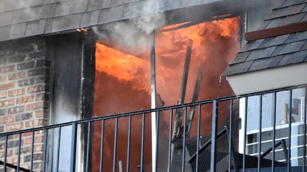 Balcony doorway on fire with bright flames and rising smoke, metal railing in front and brick wall to the left.