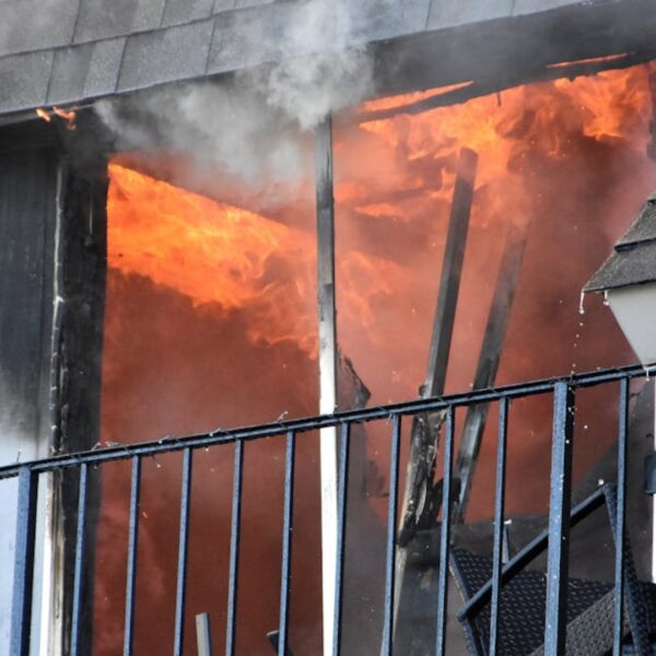 Balcony doorway on fire with bright flames and rising smoke, metal railing in front and brick wall to the left.