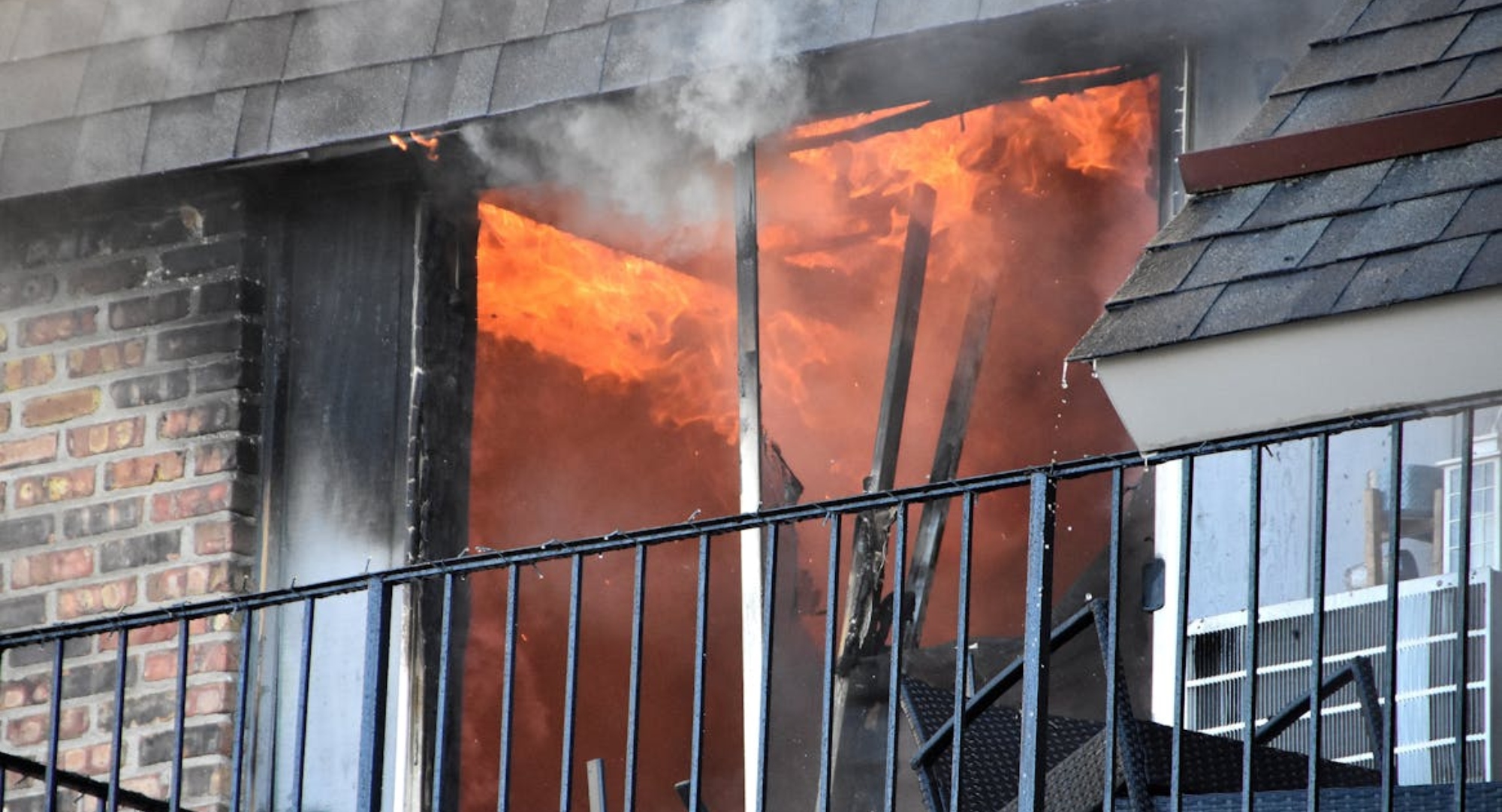 Balcony doorway on fire with bright flames and rising smoke, metal railing in front and brick wall to the left.