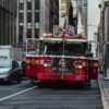 Red fire truck with 'Pride of Midtown' on the front drives down a busy NYC street, surrounded by tall buildings and yellow cabs.