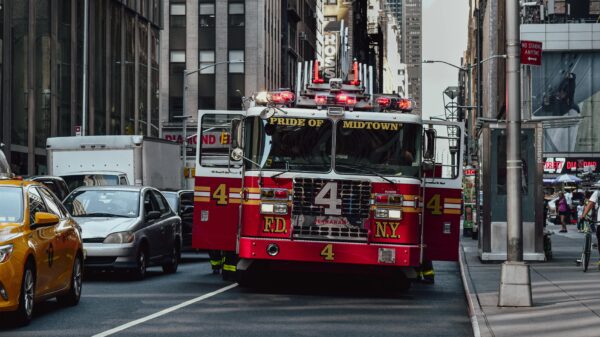 Red fire truck with 'Pride of Midtown' on the front drives down a busy NYC street, surrounded by tall buildings and yellow cabs.