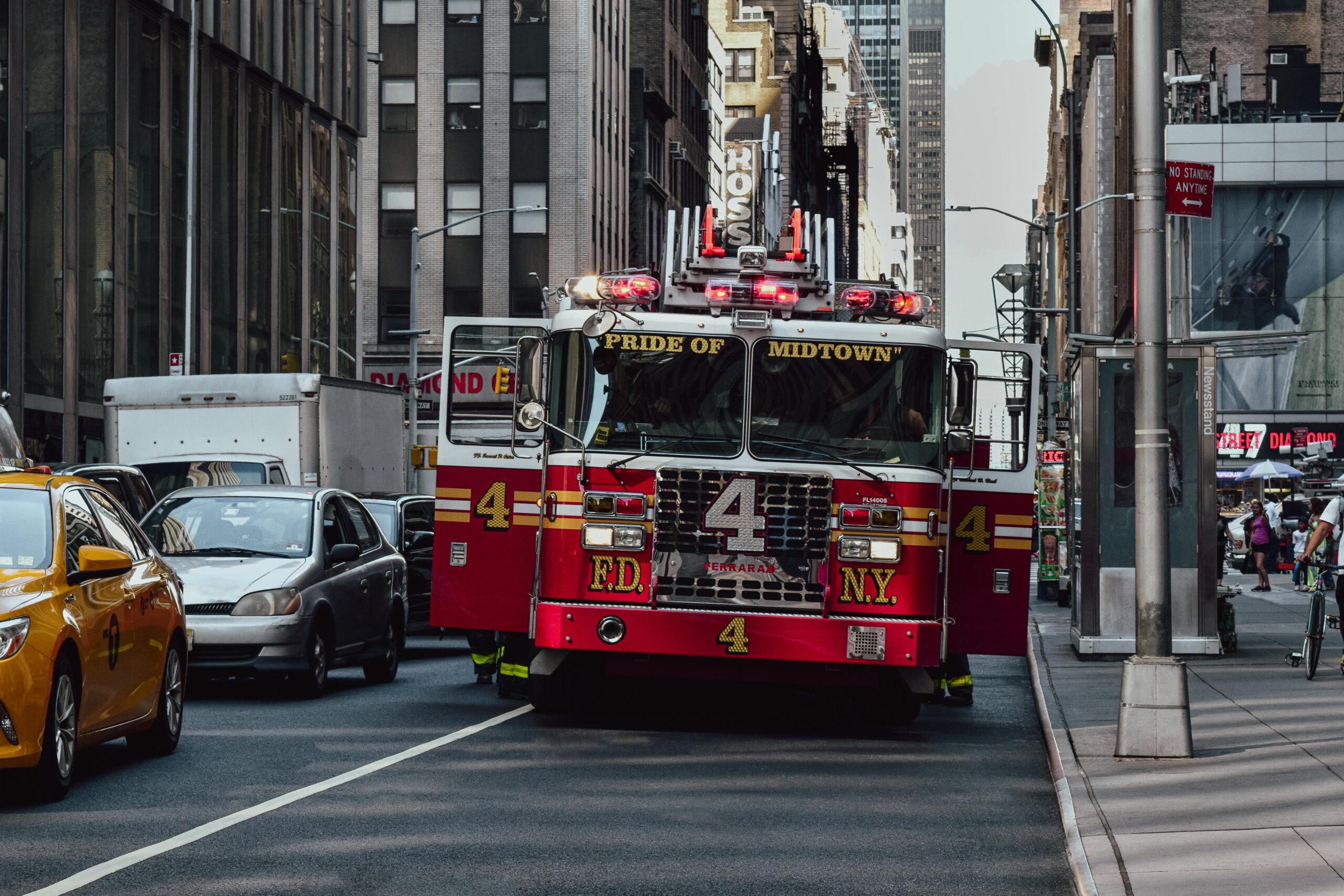 Red fire truck with 'Pride of Midtown' on the front drives down a busy NYC street, surrounded by tall buildings and yellow cabs.
