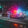 Police car with red and blue flashing lights in a snowy urban intersection at night, other vehicle nearby under snowy streetlights
