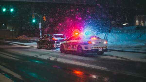 Police car with red and blue flashing lights in a snowy urban intersection at night, other vehicle nearby under snowy streetlights