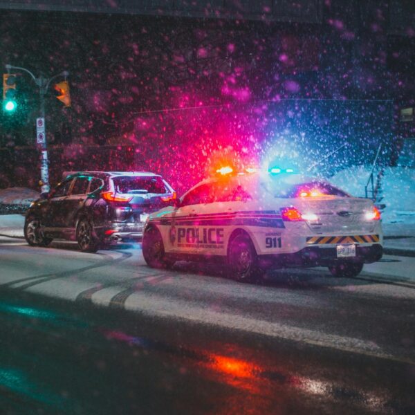 Police car with red and blue flashing lights in a snowy urban intersection at night, other vehicle nearby under snowy streetlights