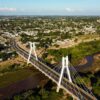 Aerial view of a white cable-stayed bridge crossing a brown river, surrounded by a green suburban landscape and city beyond.