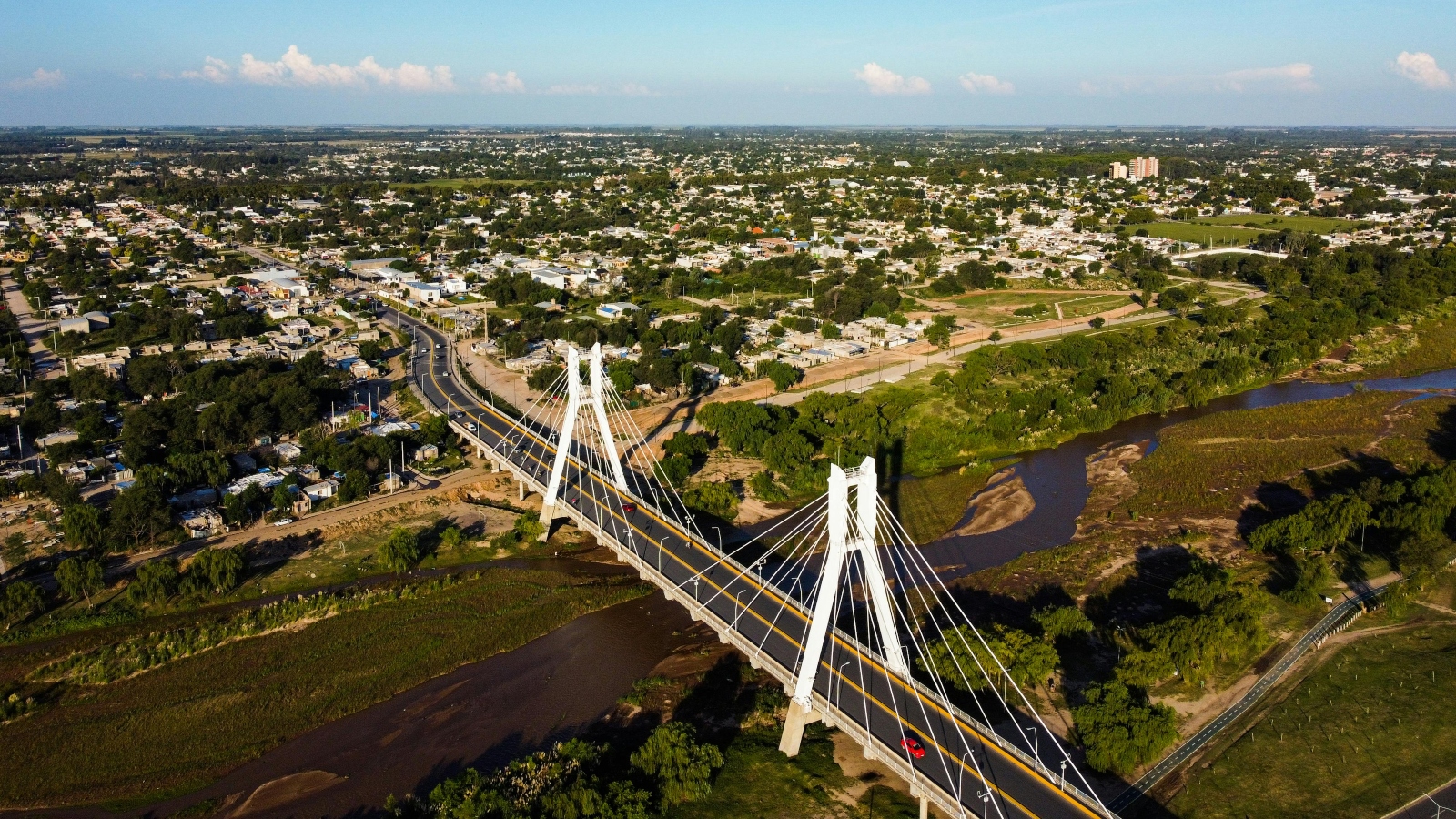 Aerial view of a white cable-stayed bridge crossing a brown river, surrounded by a green suburban landscape and city beyond.